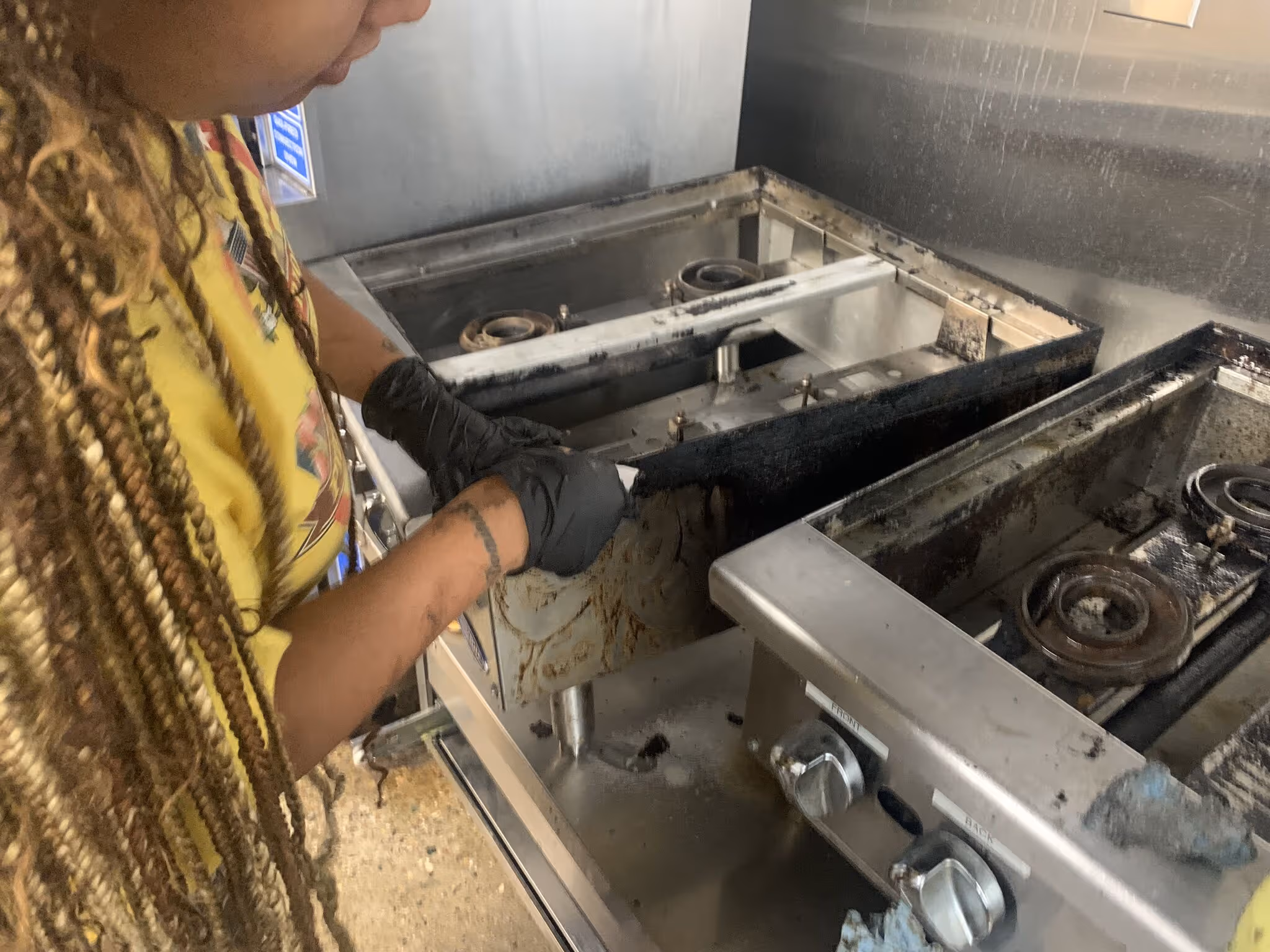 Person wearing black gloves cleaning a greasy stove burner in a kitchen.