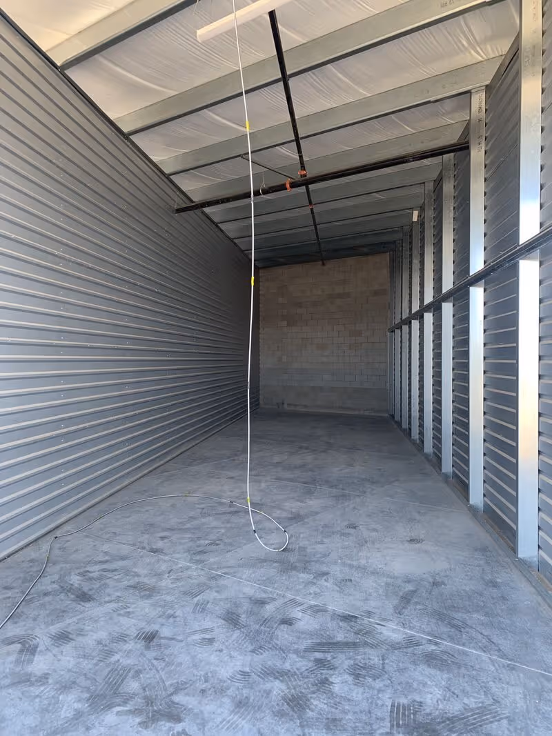 Empty industrial storage room with metal walls, concrete floor, ceiling beams, and a hanging white cable.