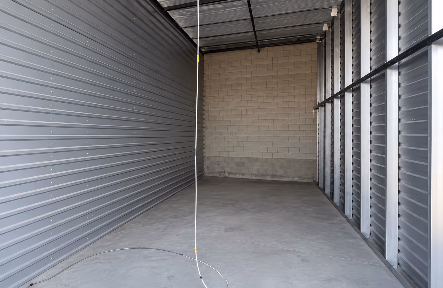 Empty storage unit with concrete floor, metal walls, and a hanging white cable in the center.