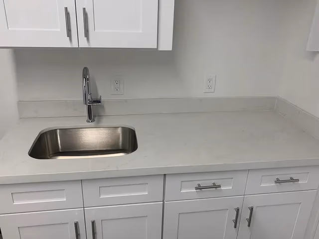 White kitchen countertop with stainless steel sink, chrome faucet, and white cabinets below and above.