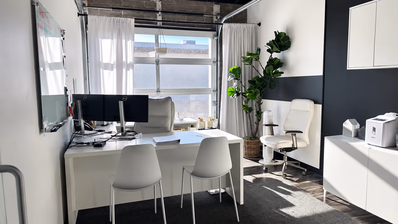 Modern office with white desks, two monitors, white chairs, a plant, and large window with curtains.
